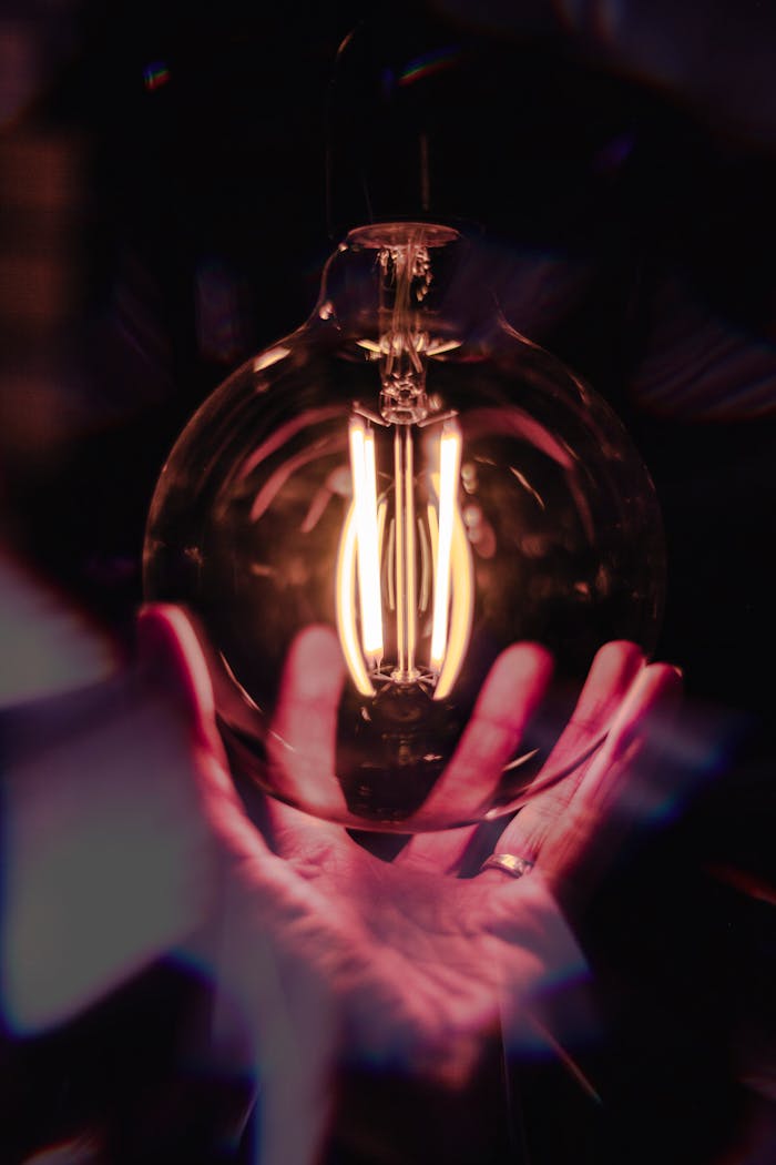 A warm glowing filament light bulb held gently in hands, creating a dramatic and artistic effect.